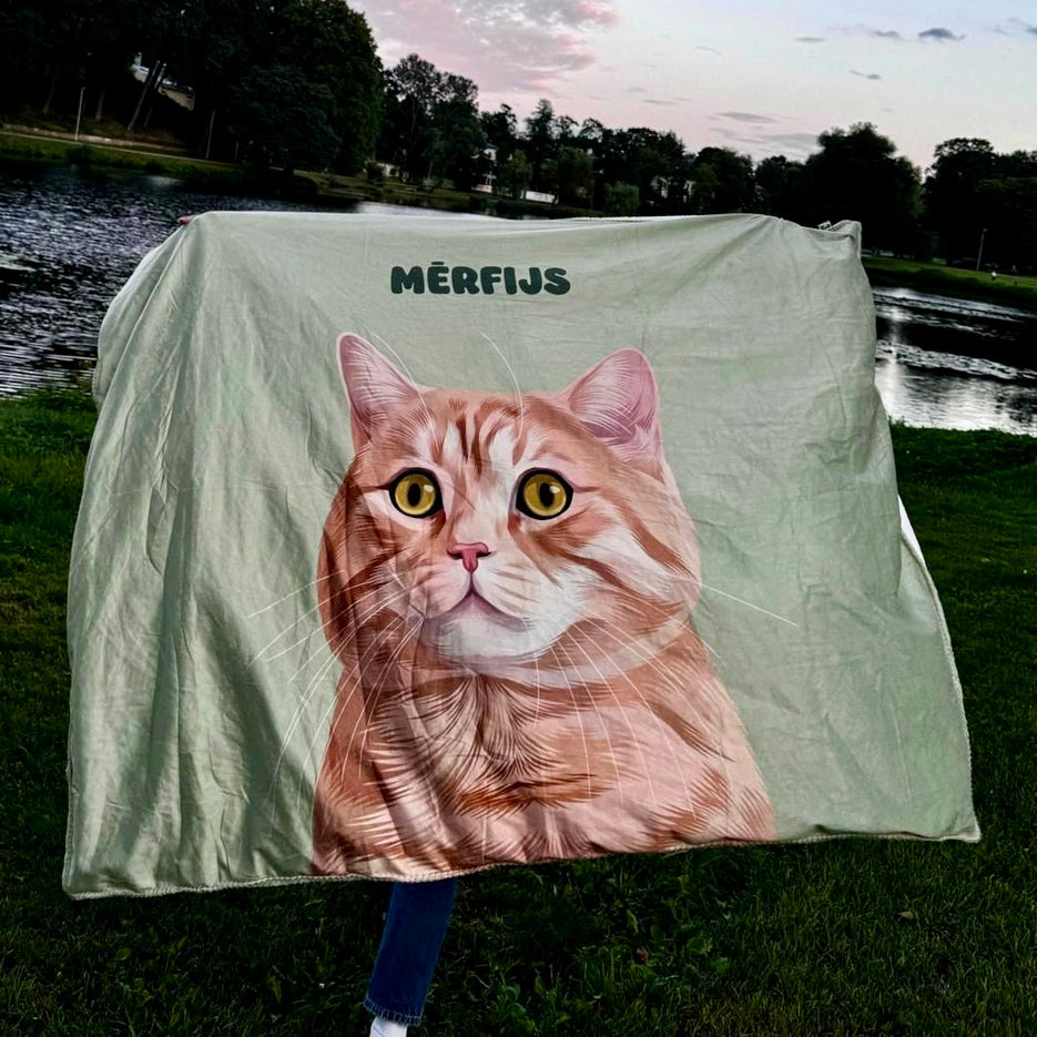 A lightweight blanket featuring a detailed illustration of a tabby cat named "Mērfijs" with striking yellow eyes, set against a soft green background, displayed outdoors near a lake with trees and a sunset sky in the background.