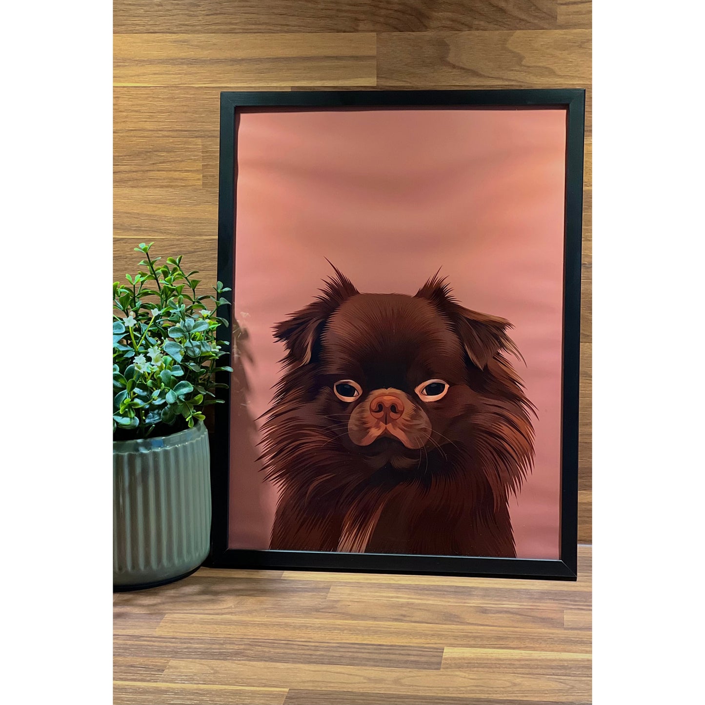 A framed wall art print featuring an illustrated image of a fluffy brown dog with expressive eyes, set against a dusty pink background, displayed on a wooden surface next to a green potted plant.