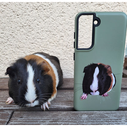 Tricolor guinea pig sitting next to a green custom pet portrait tpugh phone case featuring a custom cartoon-style portrait of the same guinea pig, placed on a wooden surface with a textured beige wall in the background.